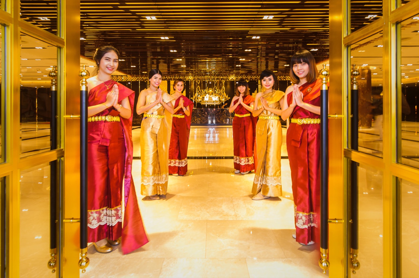 Six staff members in traditional Thai red and gold silk costumes greeting visitors with a wai gesture at the gilded entrance
